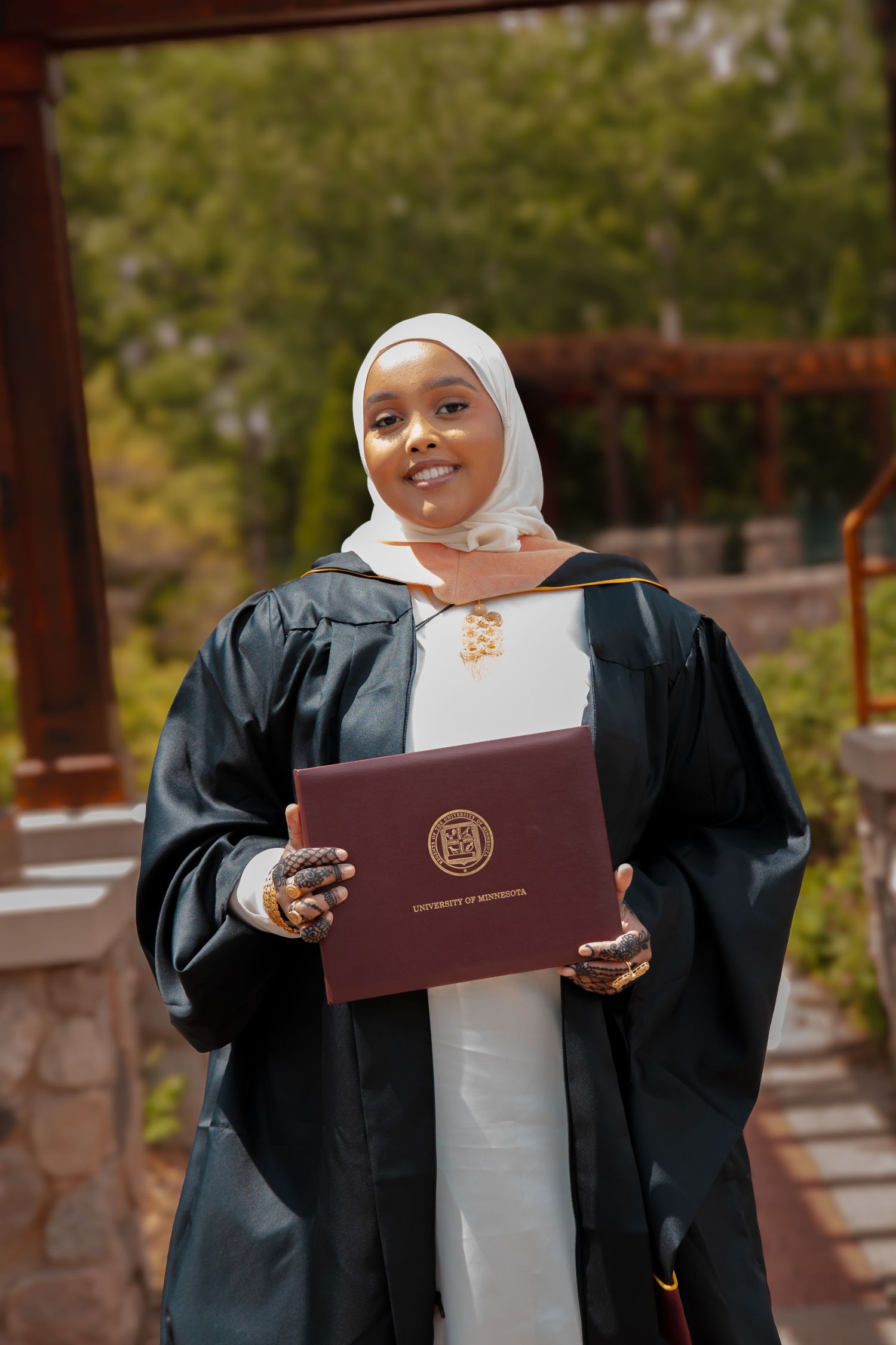 Graduate in black academic regalia holds maroon diploma holder while standing outdoors in a white hijab.
