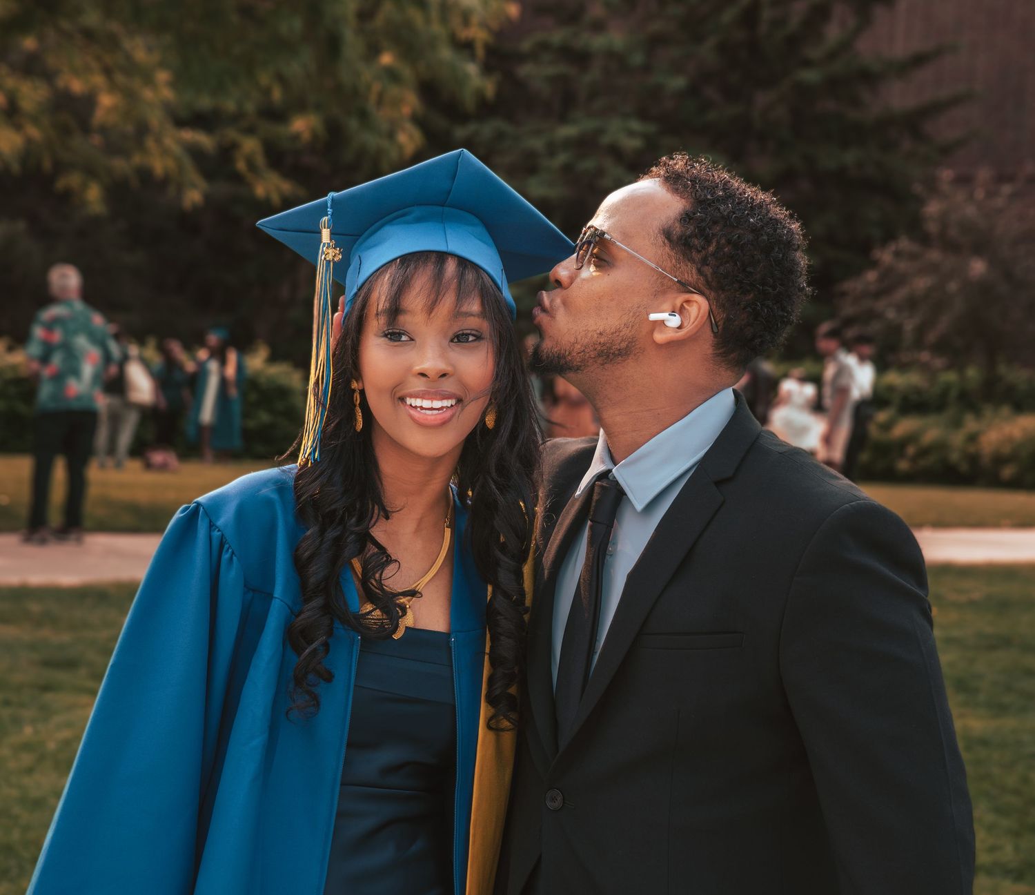 A graduate in blue cap and gown shares a tender moment with loved one during outdoor commencement celebration.