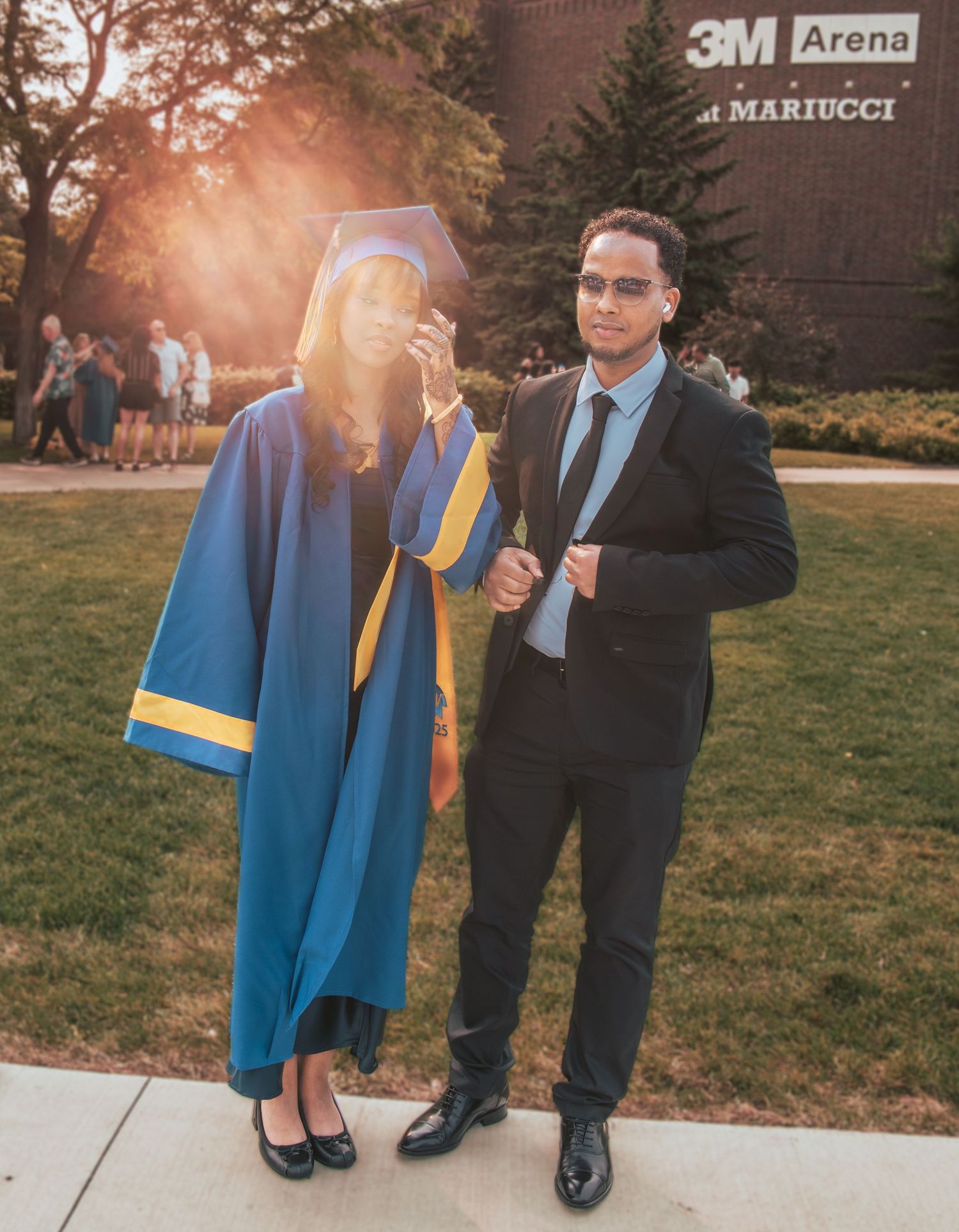 A graduation celebration photo outside the 3M Arena showing two people in formal attire during golden hour sunset.