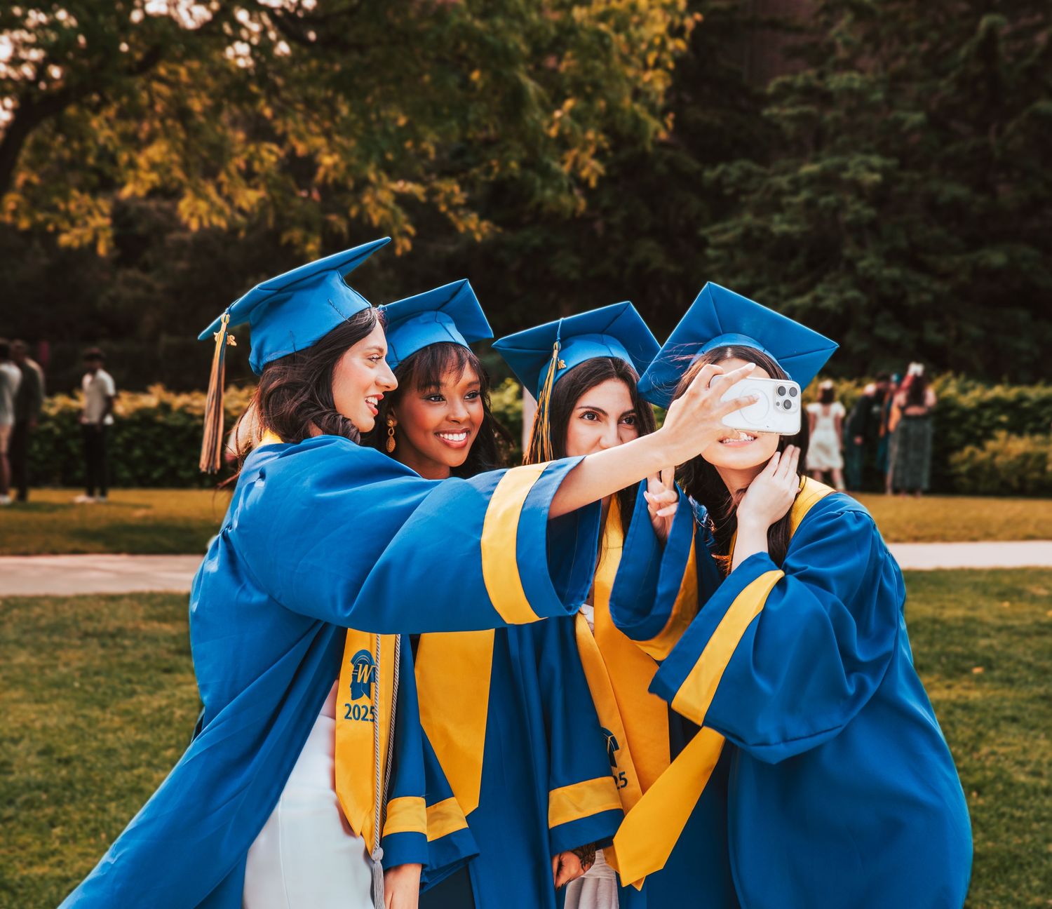 Graduates in blue and gold regalia taking a group selfie during commencement ceremony on a sunny day outdoors.