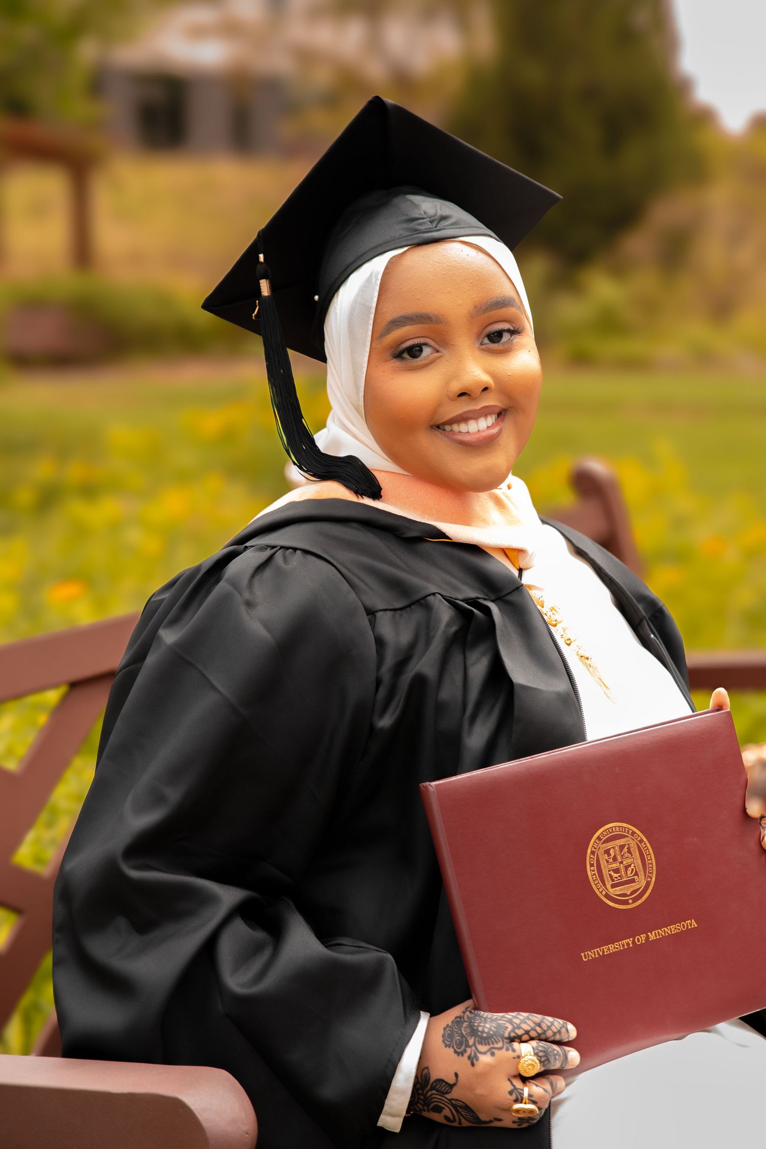 A graduate in cap and gown poses with their diploma in front of a field of yellow flowers.