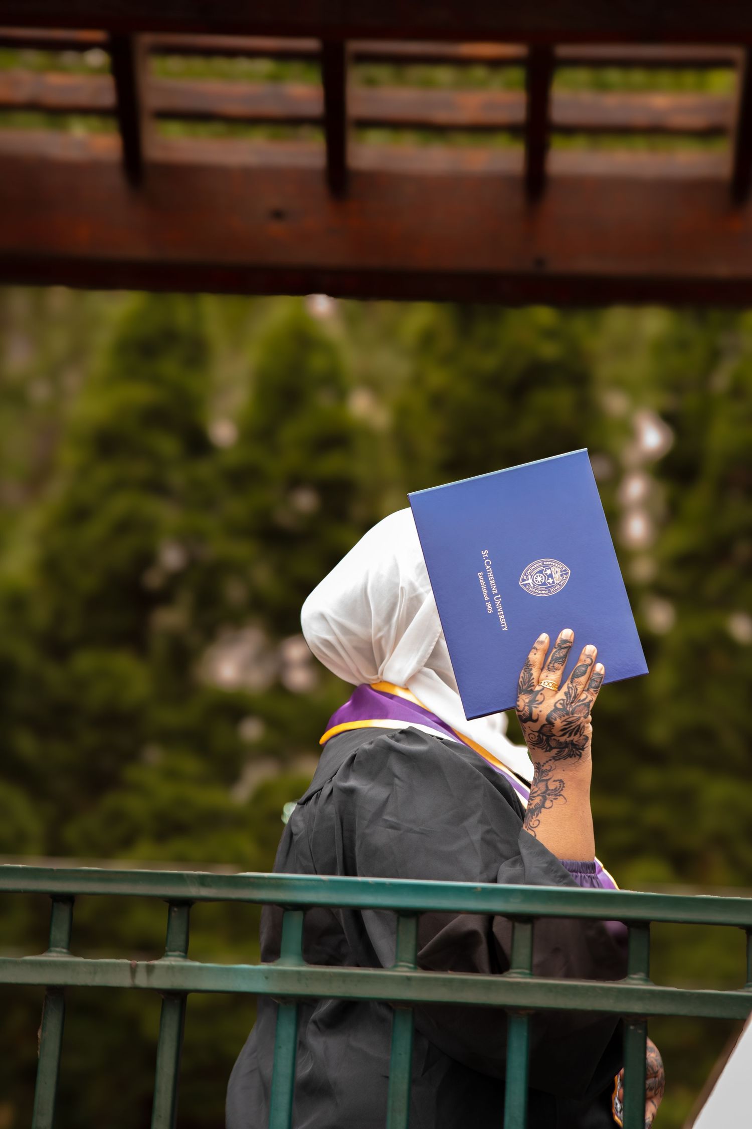 A person wearing a white headscarf holds up a blue passport or identification document while standing near a railing outdoors.