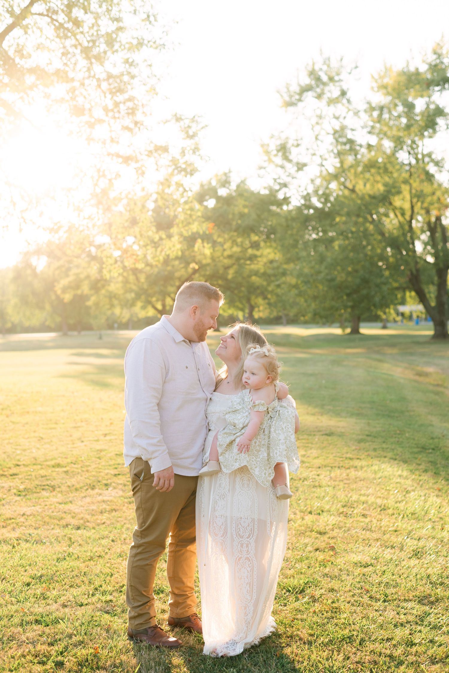 A mom and dad look lovingly at each other during golden hour while holding their toddler.
