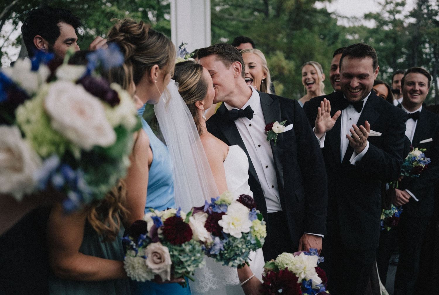 Wedding ceremony guests gather outdoors in the natural light with a mix of formal attire and floral arrangements.