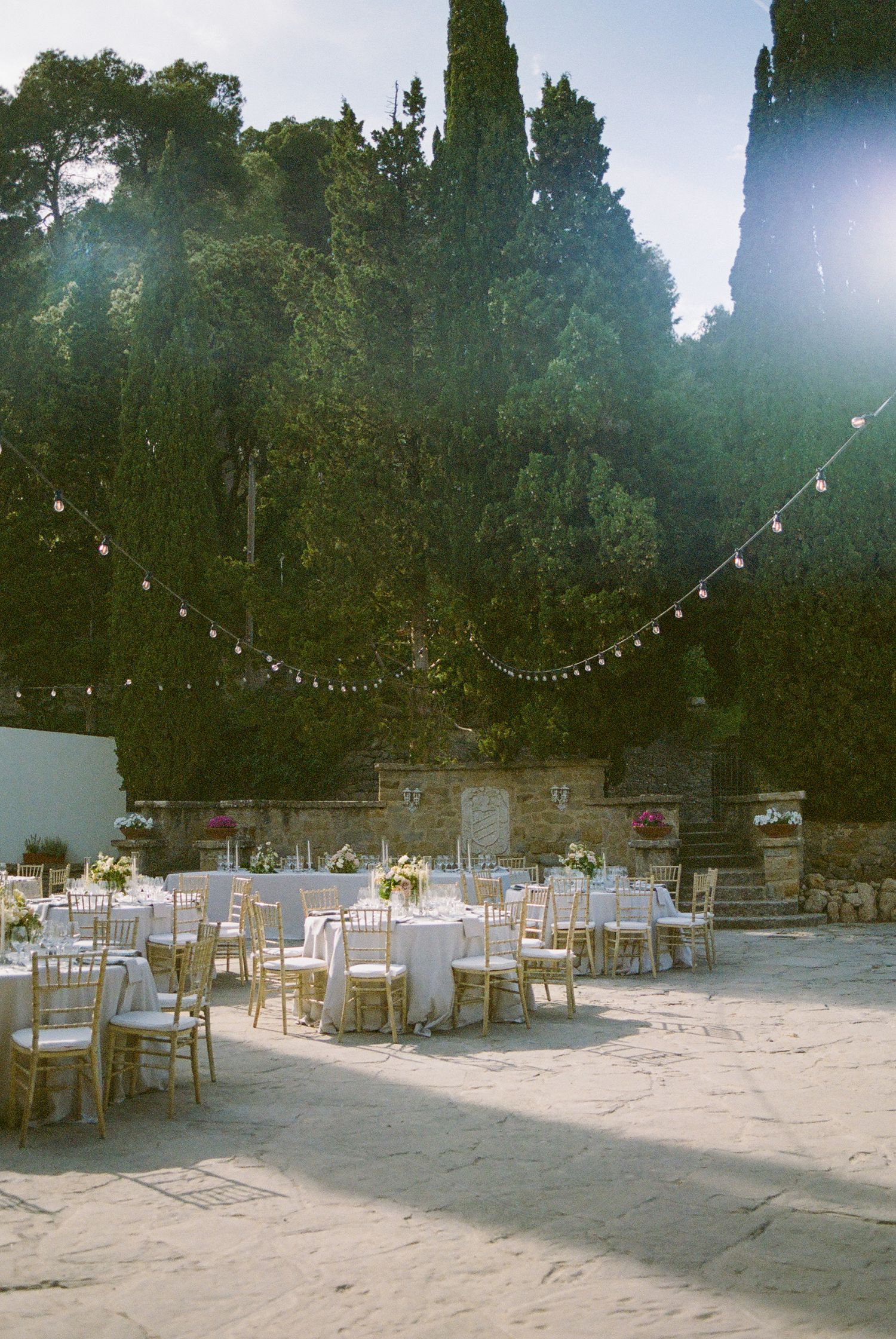 Elegant outdoor reception setup with string lights and round tables in a courtyard surrounded by cypress trees.