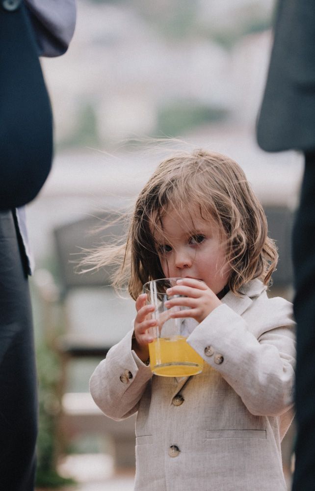 Small child in light coat sips orange juice while standing between adults.