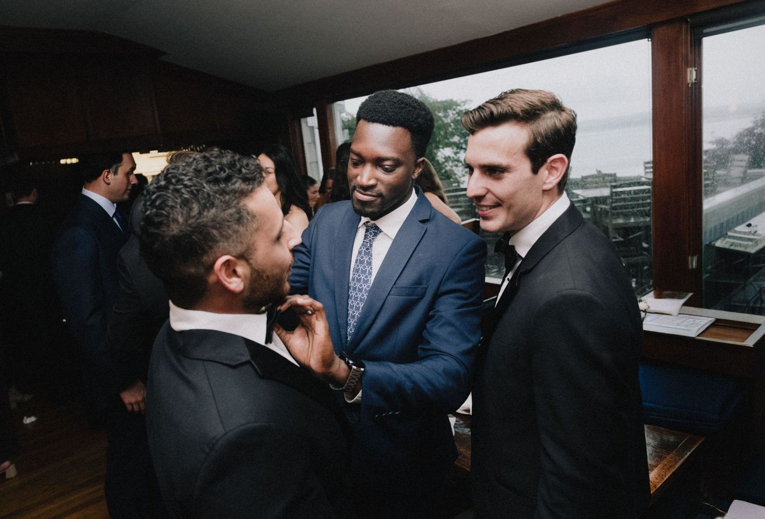 Three men wearing suits engaged in conversation near a window in a dark interior setting.
