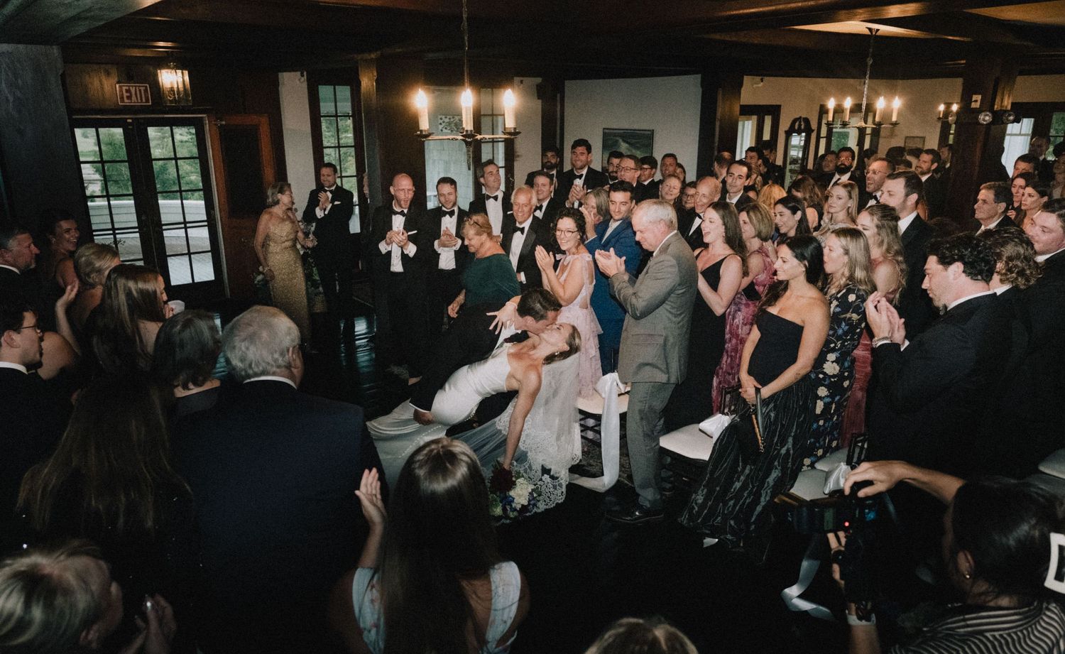 Indoor wedding reception venue filled with guests in a dimly lit rustic space with large windows.