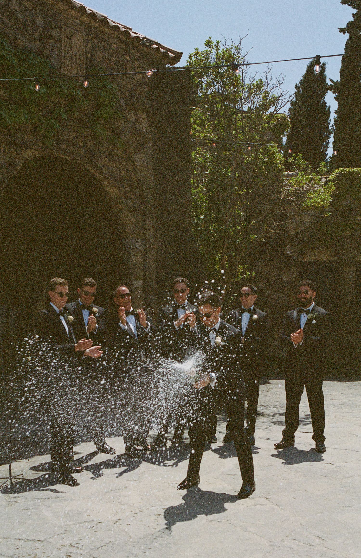 A group celebrating with champagne spray in front of a stone archway.