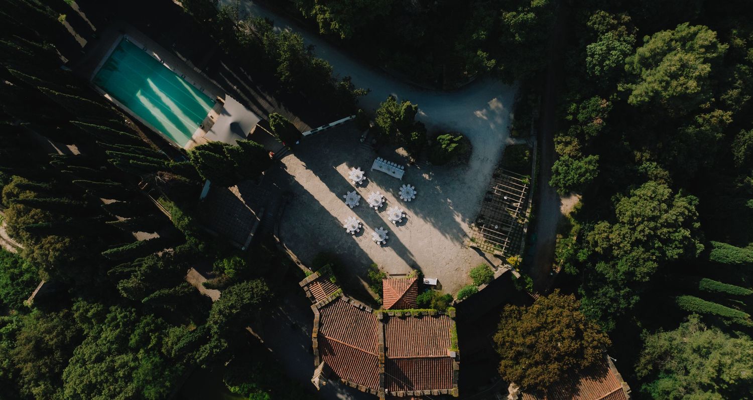 Bird's eye view of an outdoor wedding venue with pool and stone buildings surrounded by trees.
