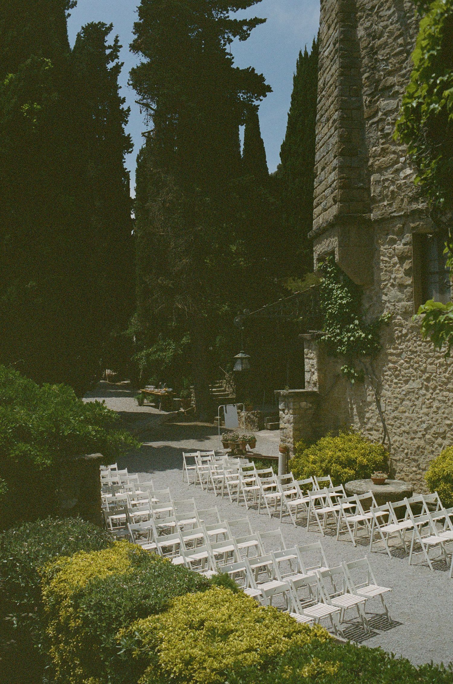 White chairs arranged for an outdoor wedding ceremony along a stone path with yellow flowers and cypress trees.