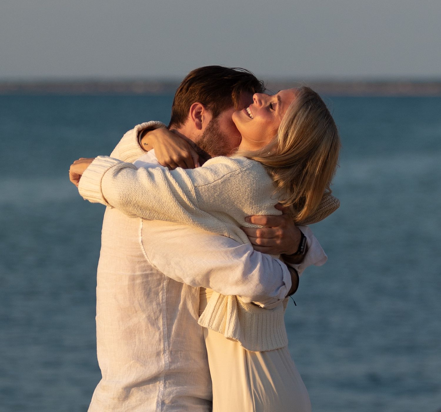 A couple embracing at sunset by the ocean wearing cozy white sweaters.