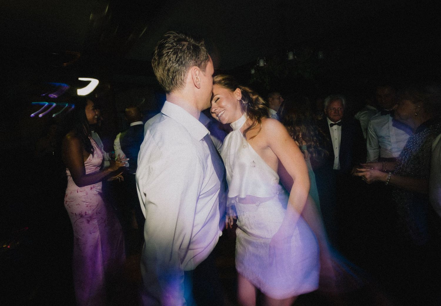 A couple shares a romantic dance at a wedding reception with purple lighting and guests in the background.