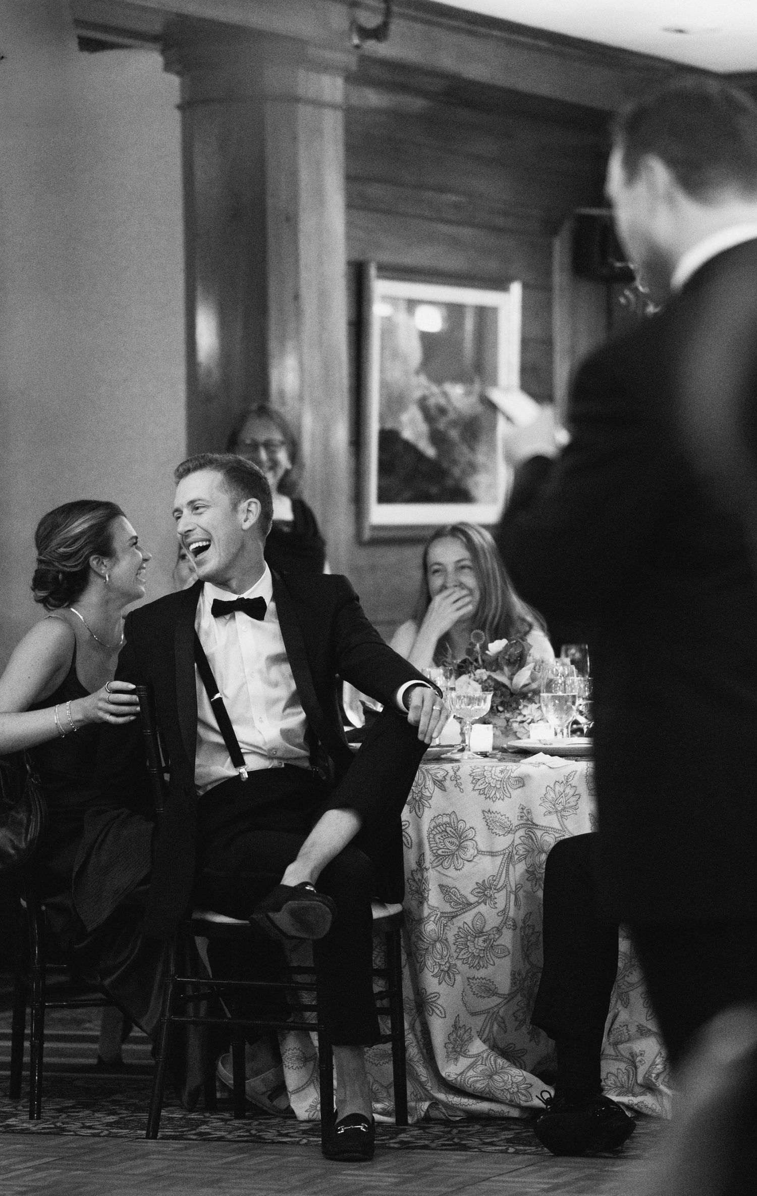 A group in formal attire shares candid laughter at an elegant black tie dinner party with subdued lighting.
