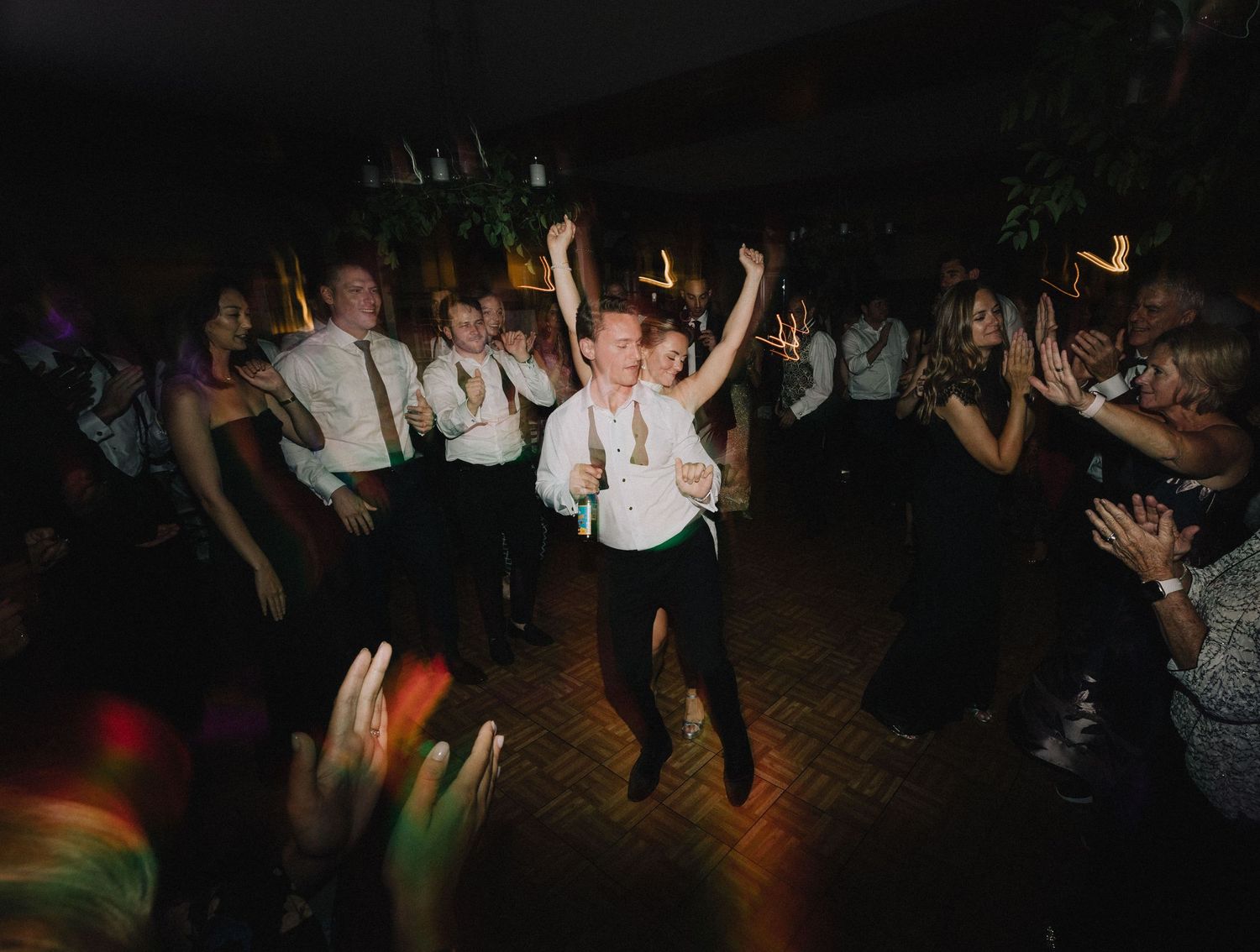 Party crowd on a dark dance floor with hands raised dancing to music under colorful lights at a nightclub event.