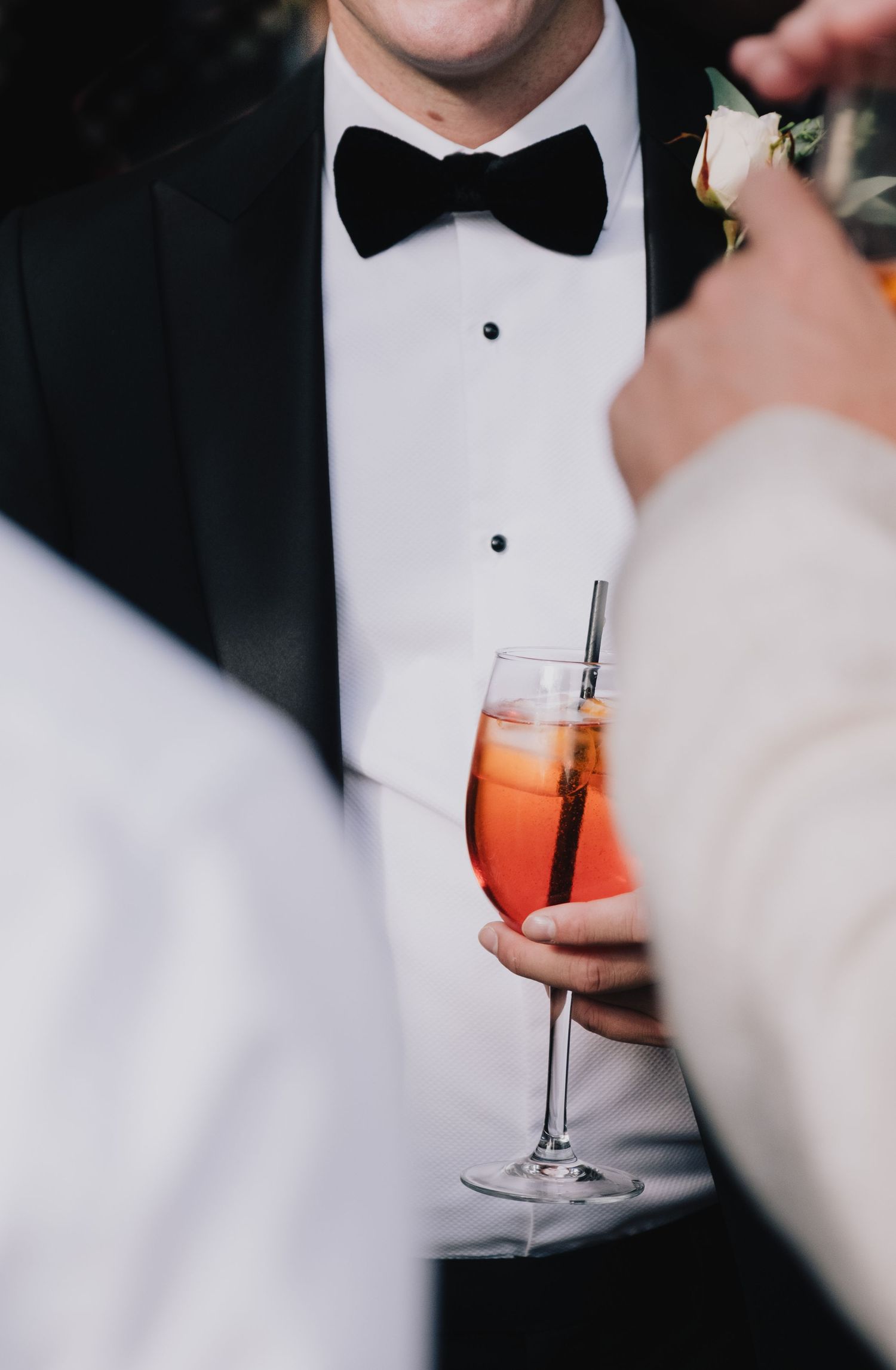 Close-up of a formal tuxedo with black bowtie and someone holding an orange cocktail.