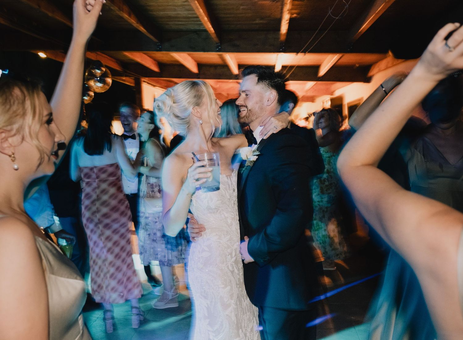 Wedding guests dancing and celebrating on a crowded dance floor with raised arms during an indoor reception party.