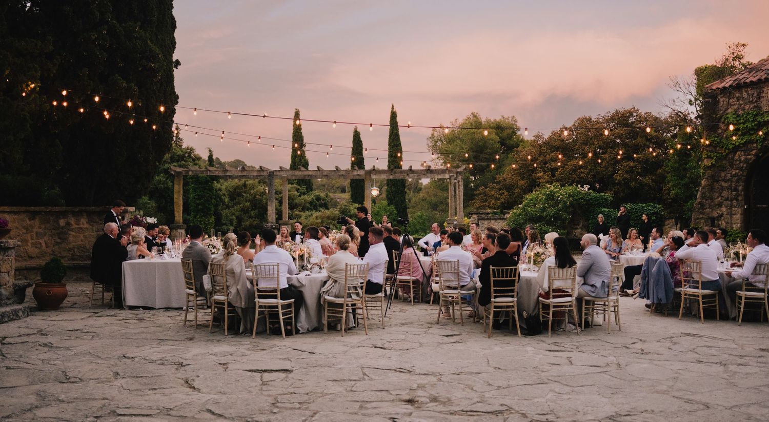 Outdoor wedding reception at sunset with string lights and cypress trees creating a romantic atmosphere.