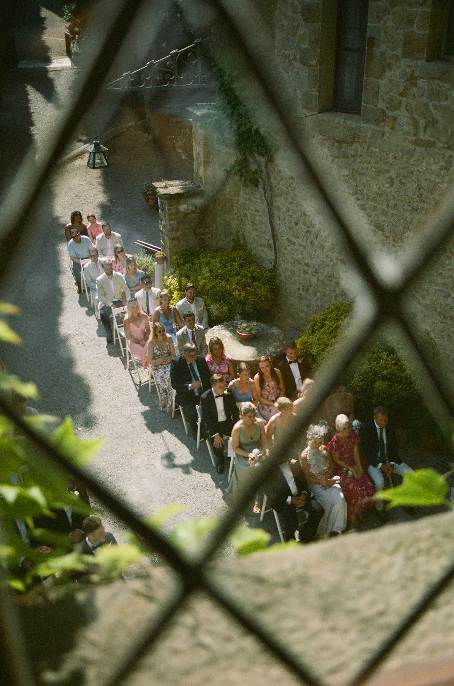 Wedding guests lined up along a stone pathway viewed through a diamond-paned window.