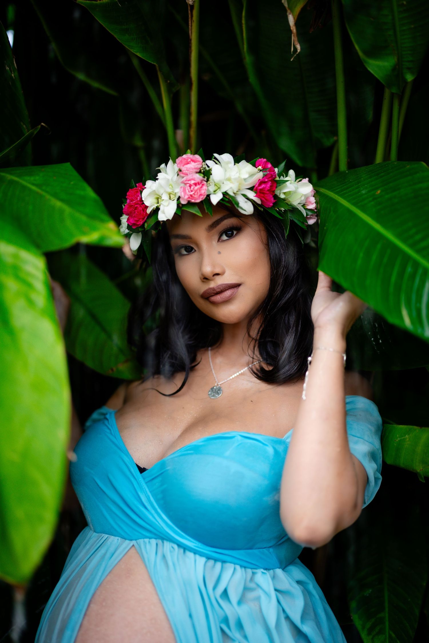 Intimate maternity portrait with a floral crown and turquoise dress surrounded by tropical leaves.