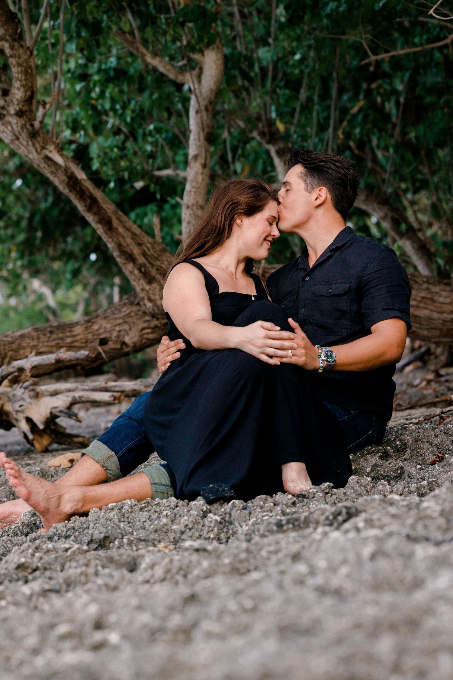 Couple sharing intimate moments while resting against a tree trunk on a beach.