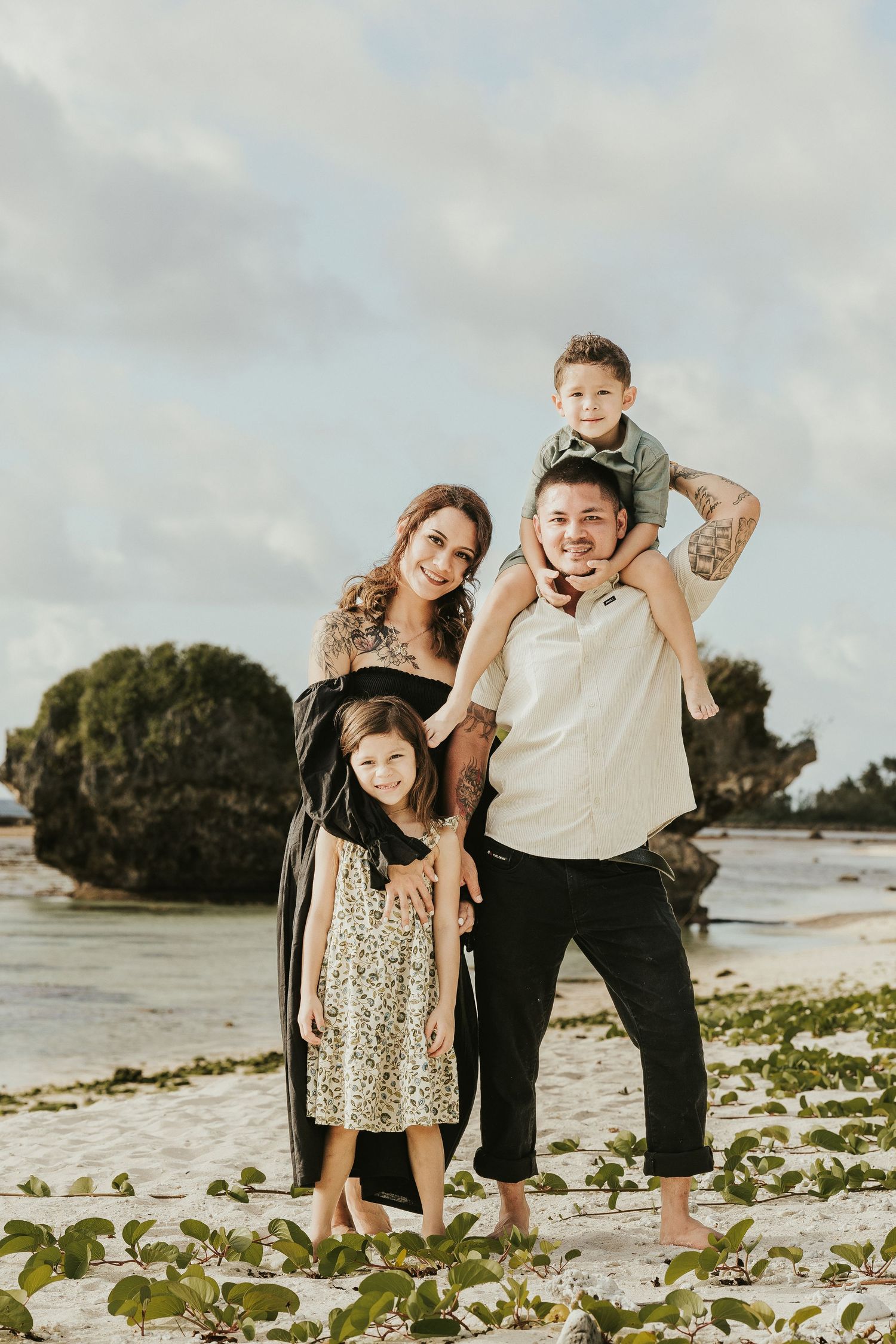 Family poses for a portrait on a beach with cloudy skies and green vegetation in the background.