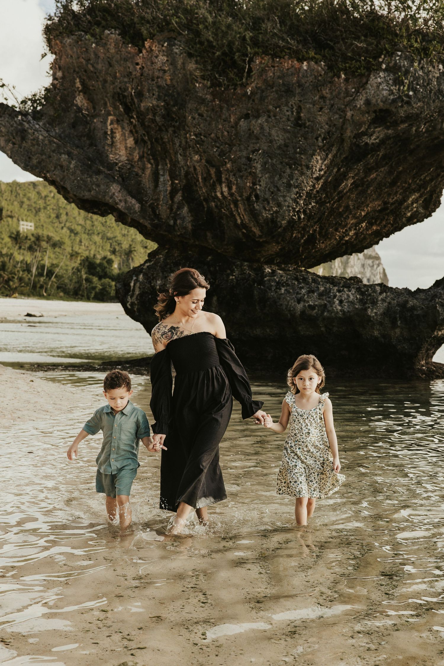 Family wading through shallow water beneath a dramatic rocky overhang on a tropical beach.