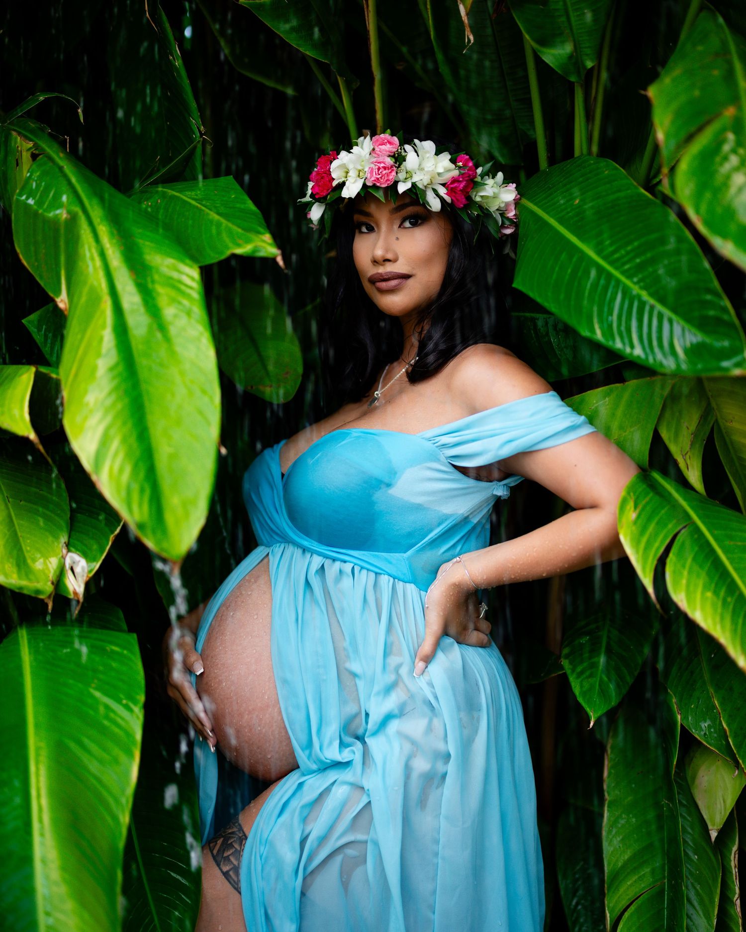 Graceful maternity photos showcasing a light blue gown and floral crown against tropical foliage.