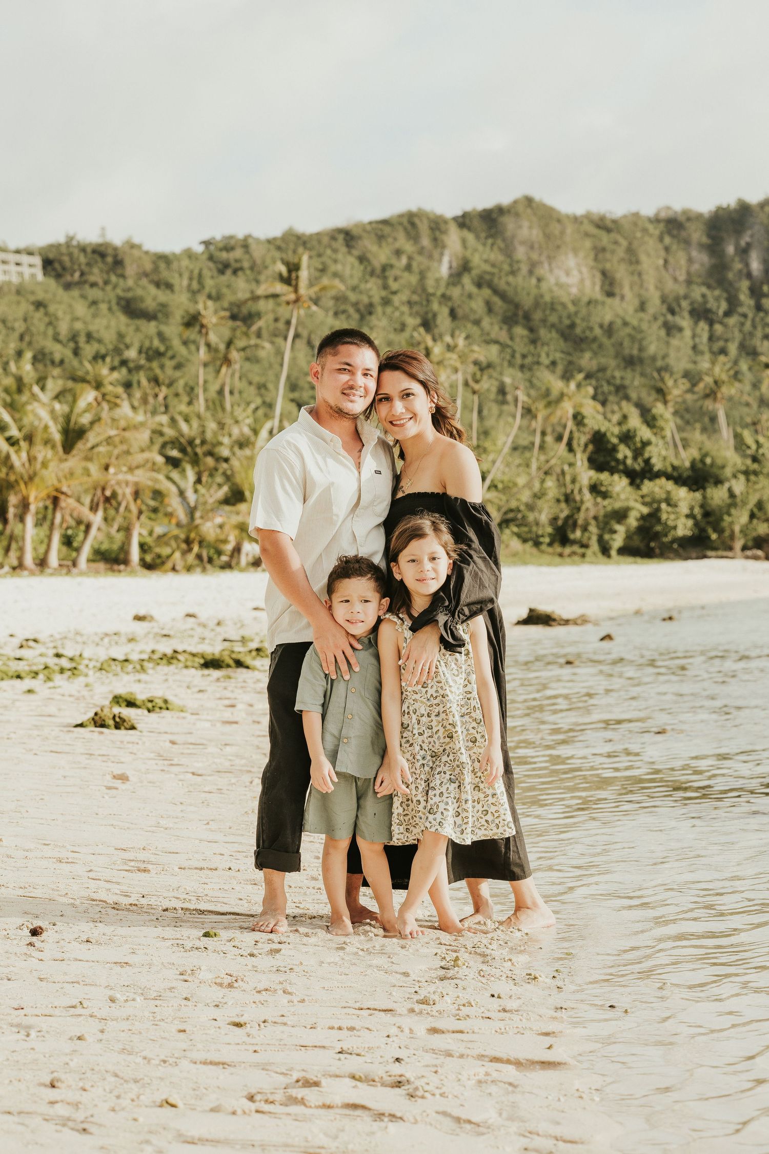 Family poses together on a sandy beach with tropical mountains in the background.