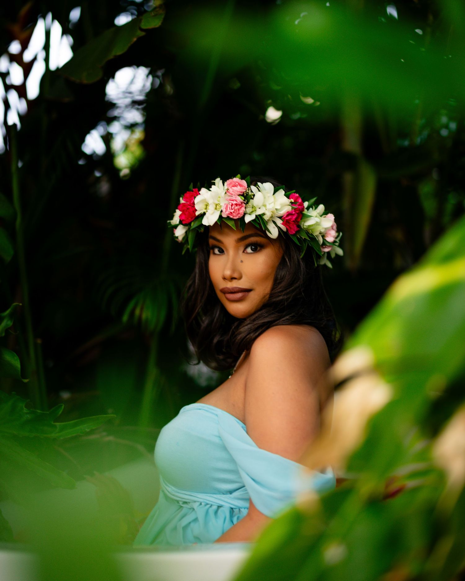 A floral crown adorns a figure in a light blue dress amid lush tropical greenery and dappled sunlight.