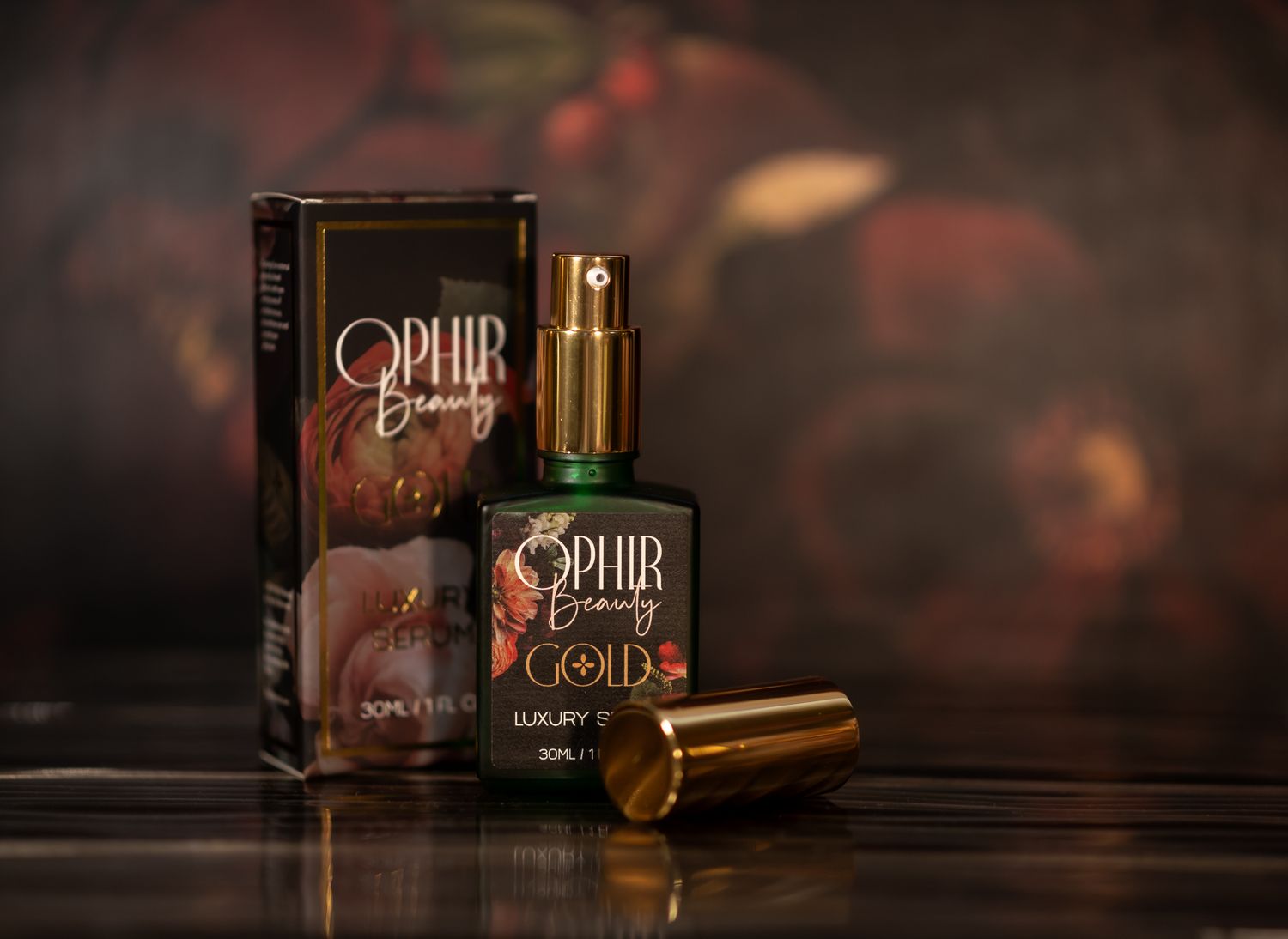 an uncapped bottle of Ophir Gold serum stands with it's gilded retail box in front of a dark floral wall, showing it's elegant pump-top.