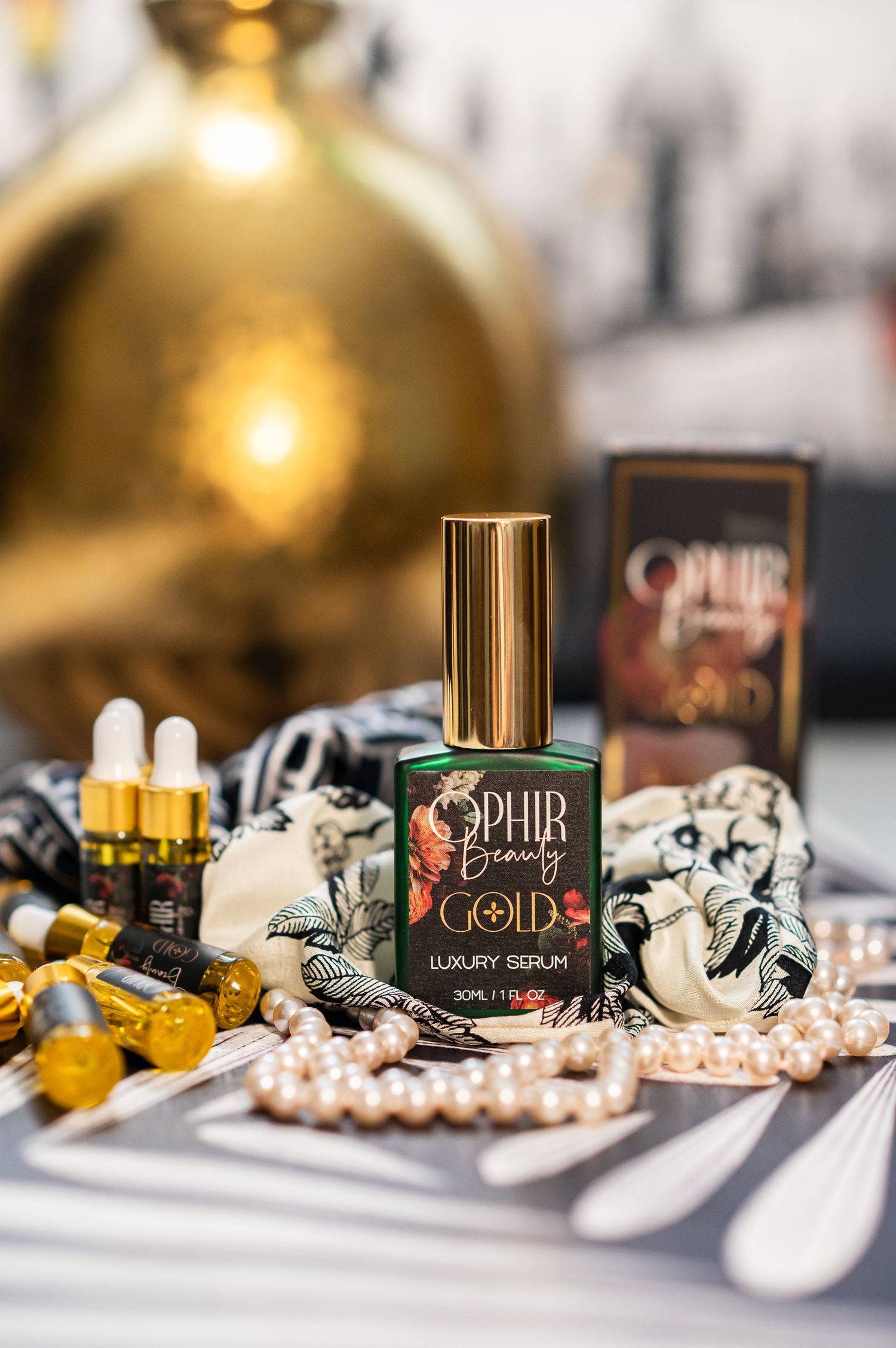a bottle of Ophir Beauty Gold serum sits in a grouping with sample sized bottles and pearls on a bold black and white surface