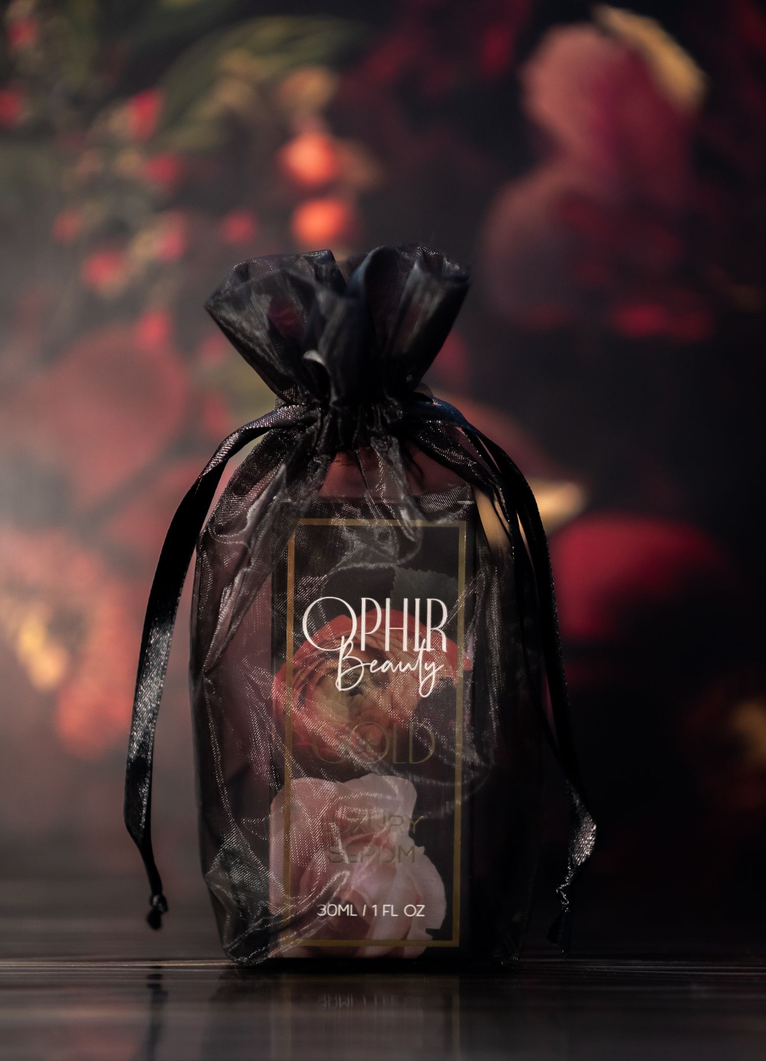a boxed Ophir Beauty Gold serum in a black organza bag sits on a dark surface with rich floral wallpaper in the background