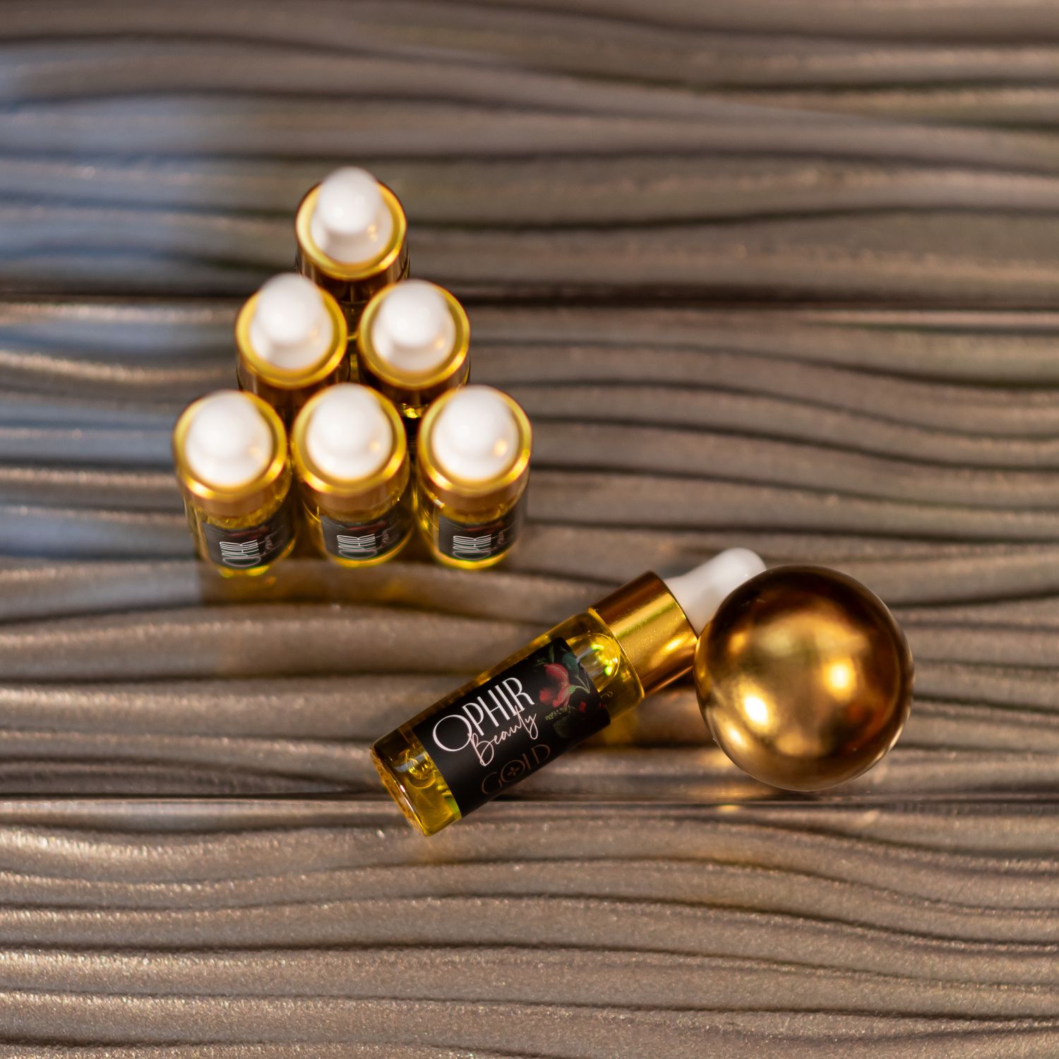 Several small sample dropper bottles of Ophir Gold serum are arranged in a pyramid, with one lying on its side, on a rippled, gold surface