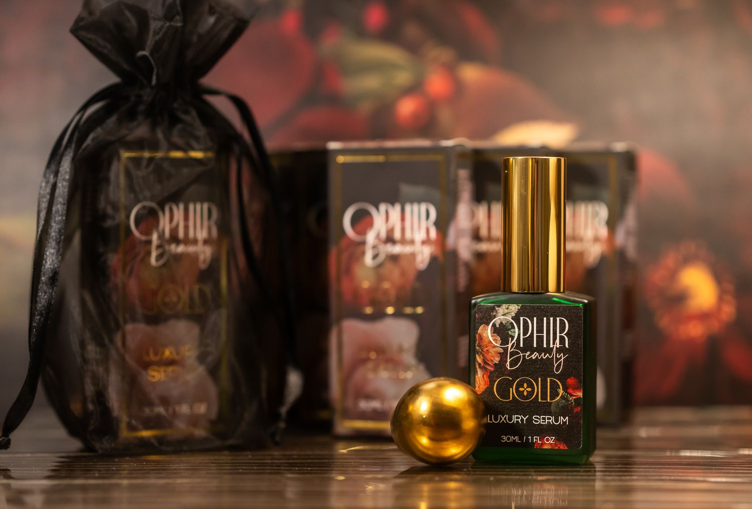 A grouping of Ophir Gold products are arranged on a reflective dark surface, with a vintage floral wallpaper in the background