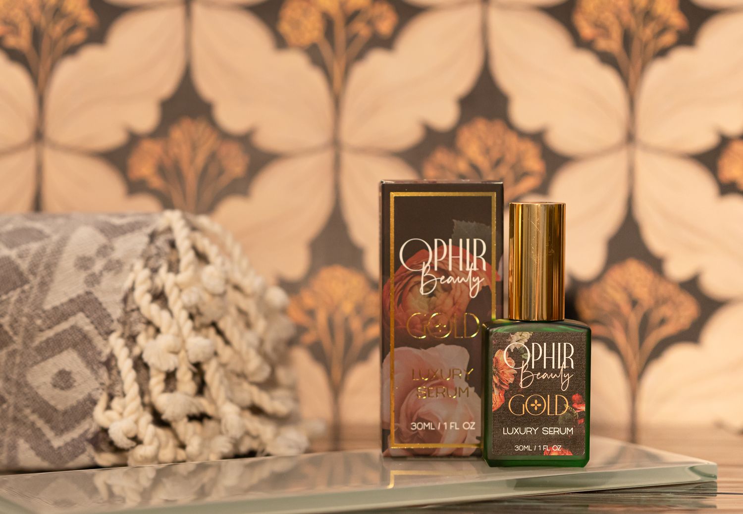 Ophir Gold luxury Serum in a green glass bottle sits with its gilded retail box on a glass tray, with a rolled up spa towel on one side