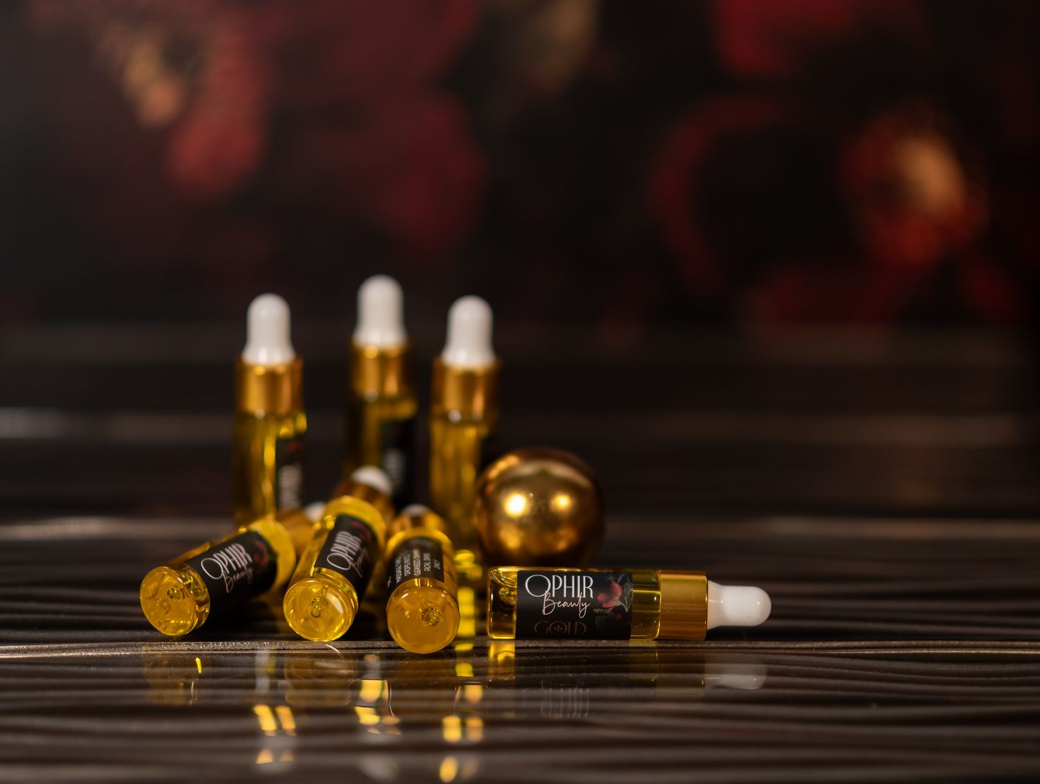 a grouping of Ophir Beauty Gold Serum sample sized dropper bottles stand and lie on a dark surface with dark florals in the background