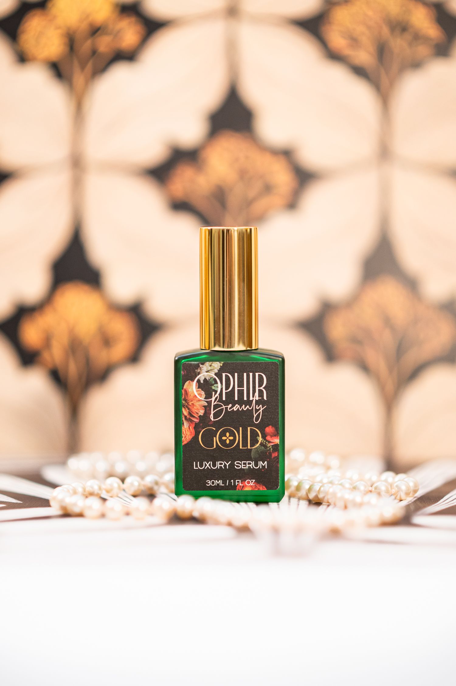 a bottle of Ophir Gold luxury serum stands on a white surface surrounded by strings of pearls, with vintage wallpaper in the background