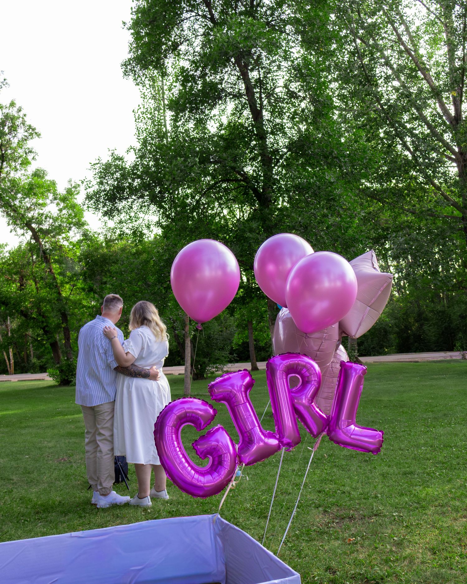 Couple embracing in a park beside pink GIRL foil letter balloons at a gender reveal celebration.