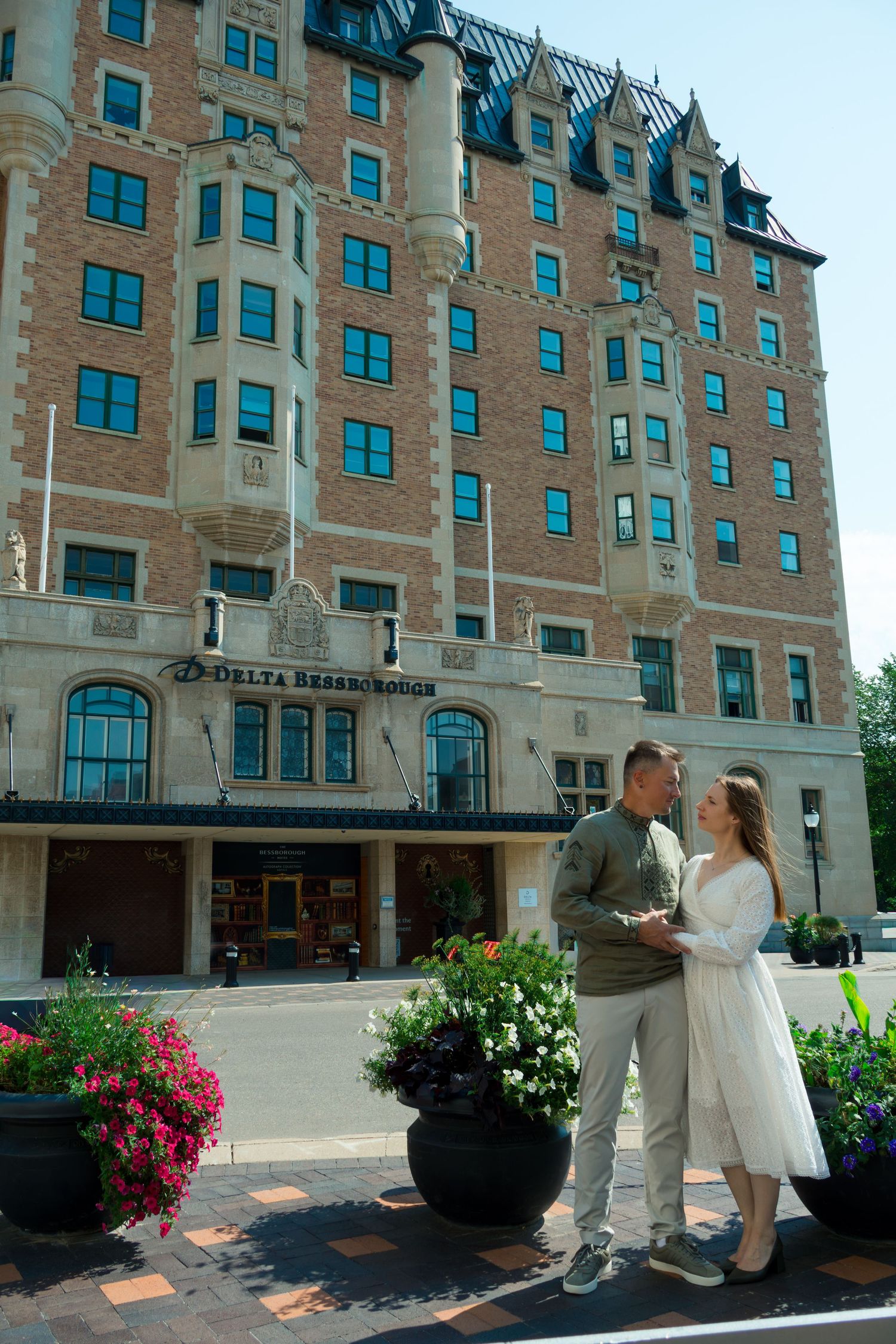 Couple embracing on a sunny street in front of the historic Delta Bessborough hotel, with colorful flower planters in the foreground.