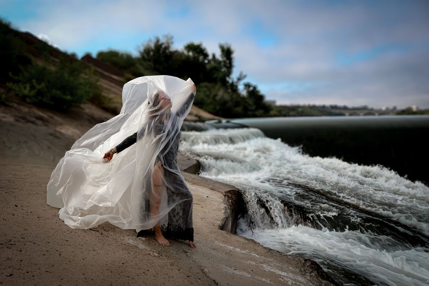 Person draped in flowing sheer white fabric stands on a sandy riverbank beside rushing white-water rapids under a soft blue sky.