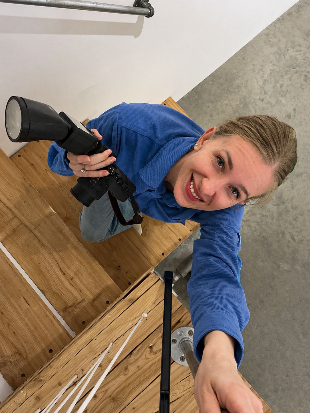 Smiling woman in a blue sweater holds a camera with a flash while climbing a wooden staircase, shot from a high angle looking down.