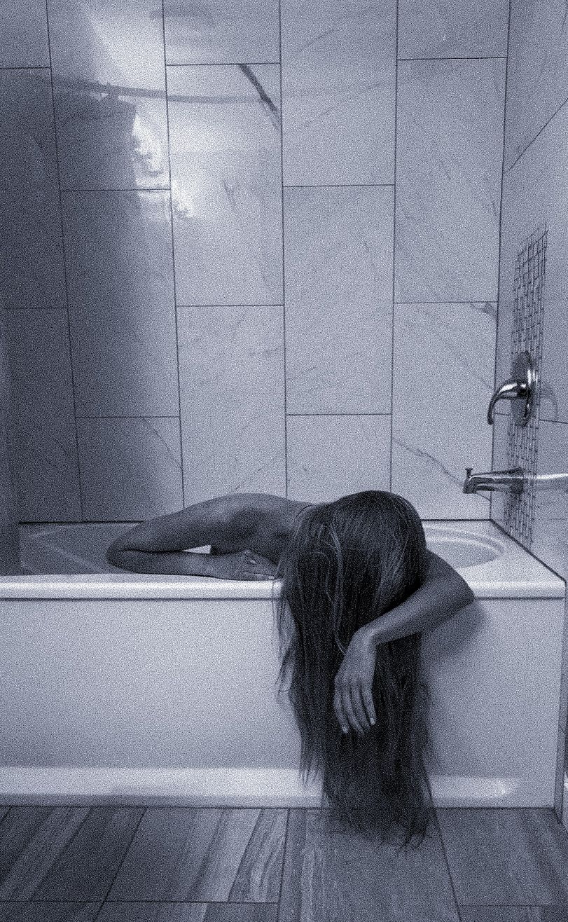 Black and white photo of a person with long hair draped over the side of a marble-tiled bathtub, one arm hanging down, in a moody scene.