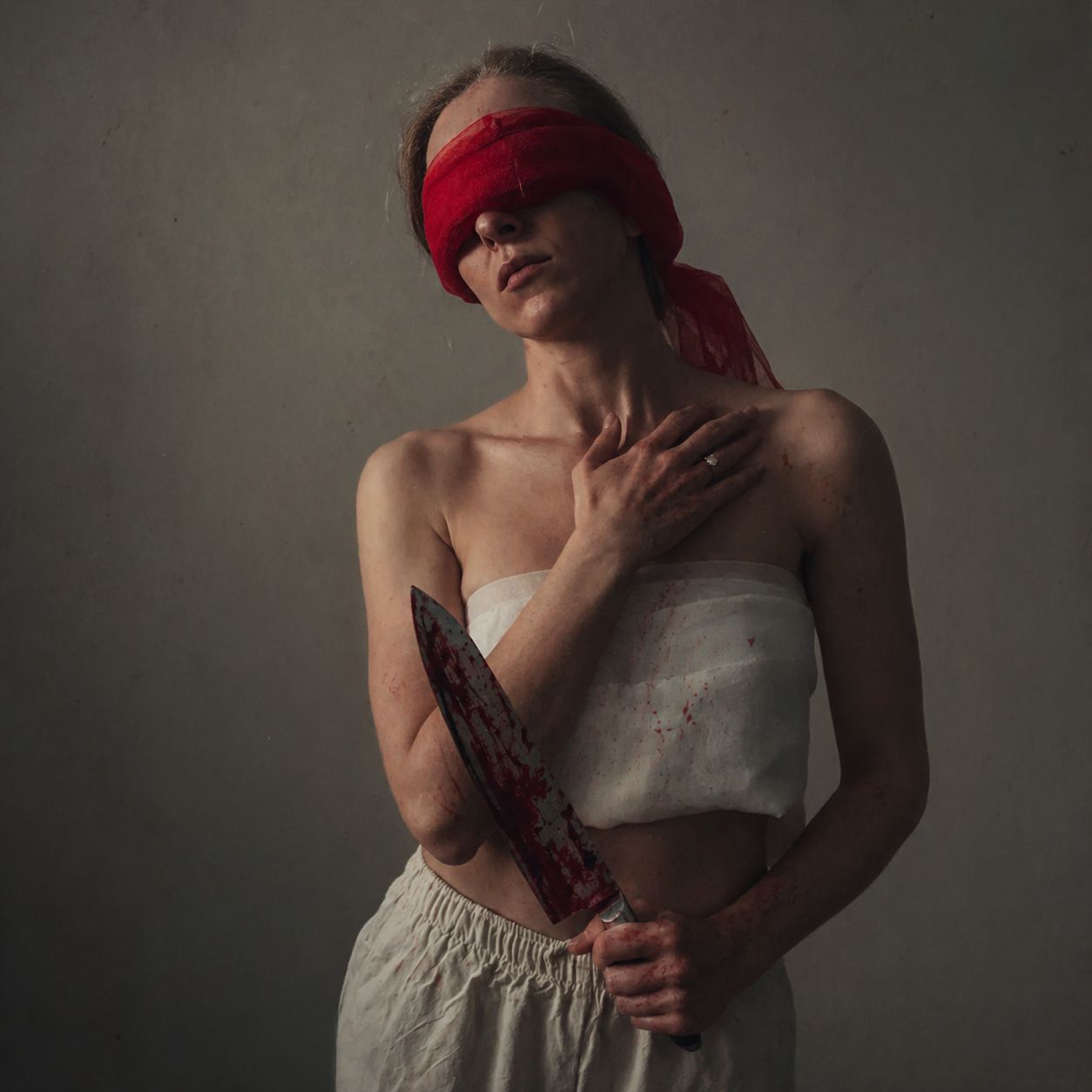 Blindfolded woman in a white top and pants holds a blood-stained knife against a muted gray wall in a dramatic, moody portrait.