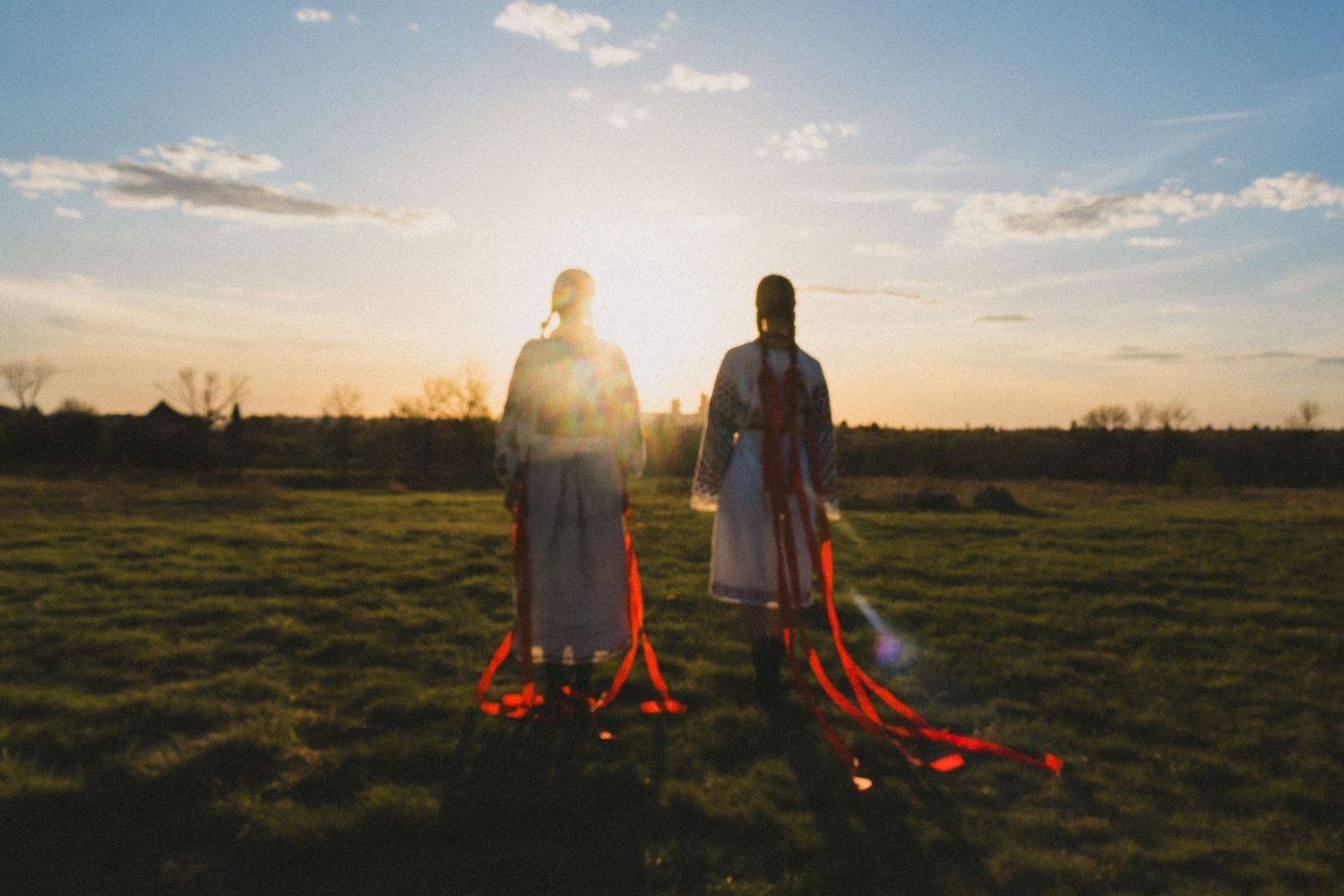 Two people in long skirts with red ribbons stand facing a glowing sunset over a grassy prairie field with bare trees in the distance.