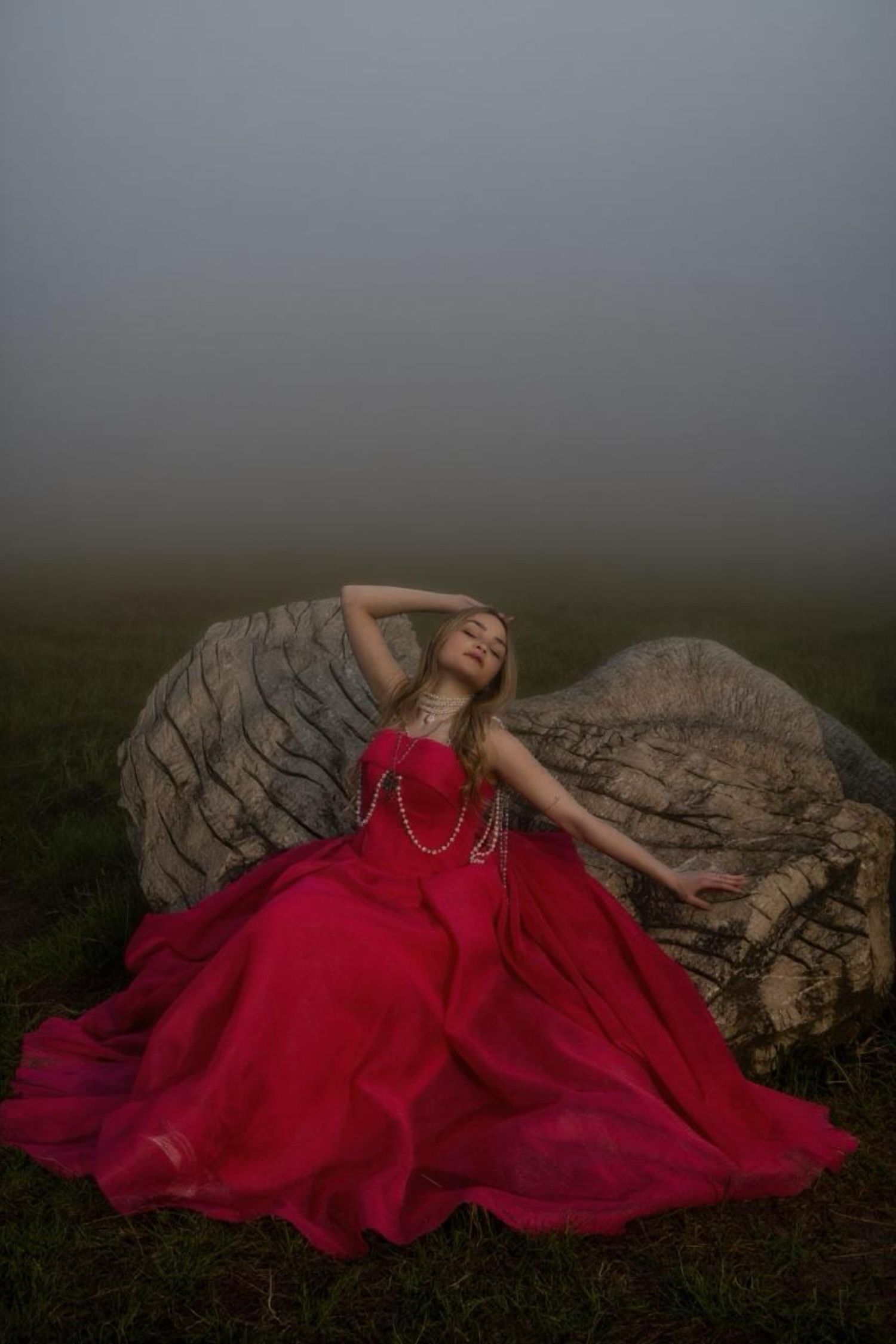 Woman in a flowing red gown and pearl necklaces reclines against a large rock in a misty, moody field.