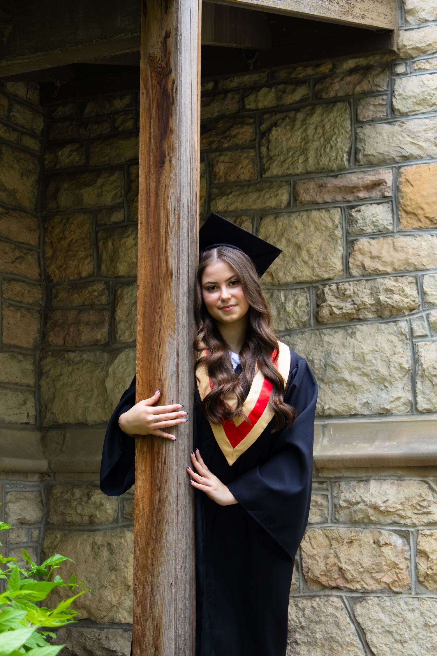 Smiling graduate in a black cap and gown with a red and gold stole poses beside a wooden post against a rustic stone wall.