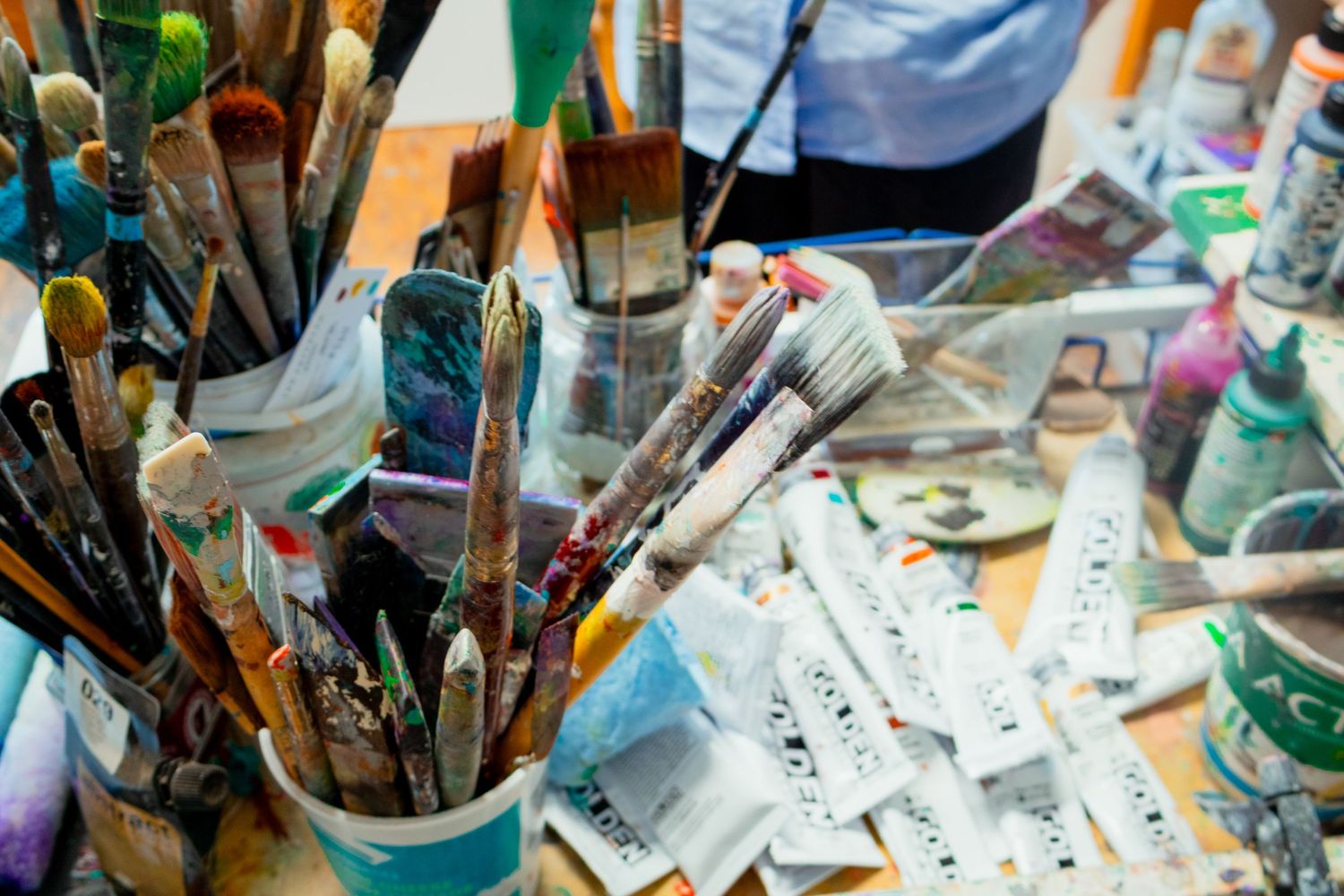 Paintbrushes in jars and tubes of paint sit on a cluttered artist's table with palette knives and labeled paper strips in a creative studio.