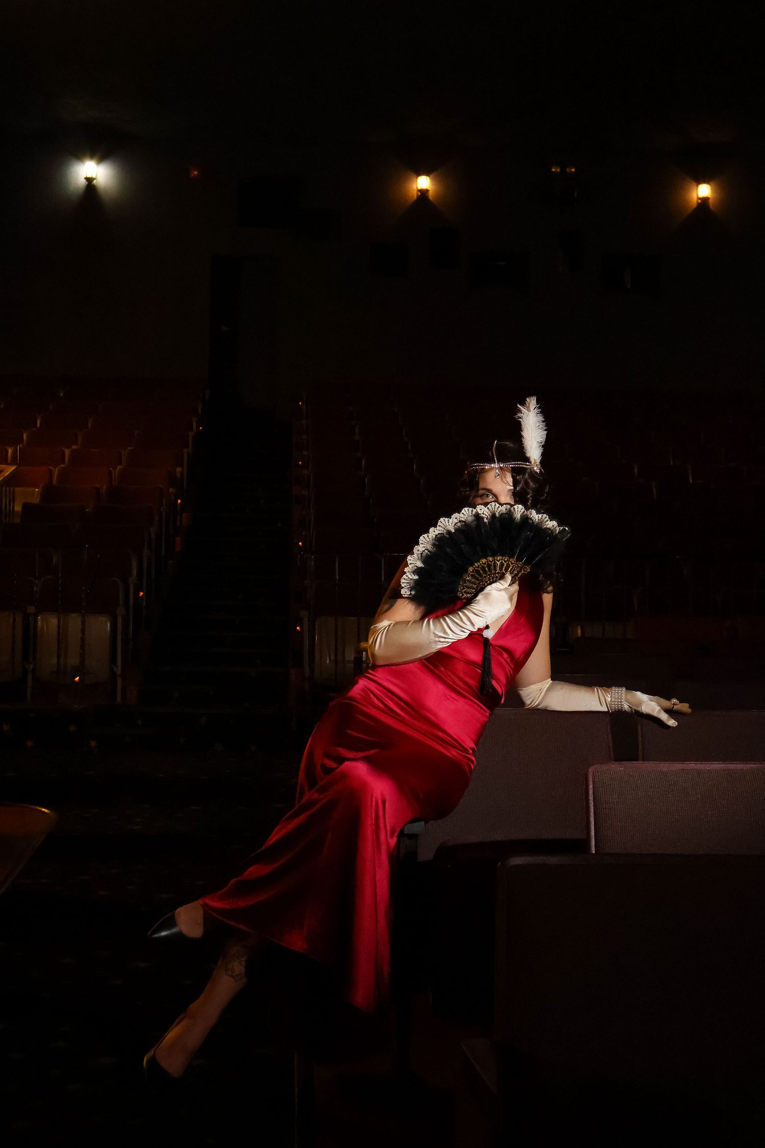 Woman in a red satin gown, white gloves and feathered headband holds a black feather fan while seated in a dimly lit vintage theater.