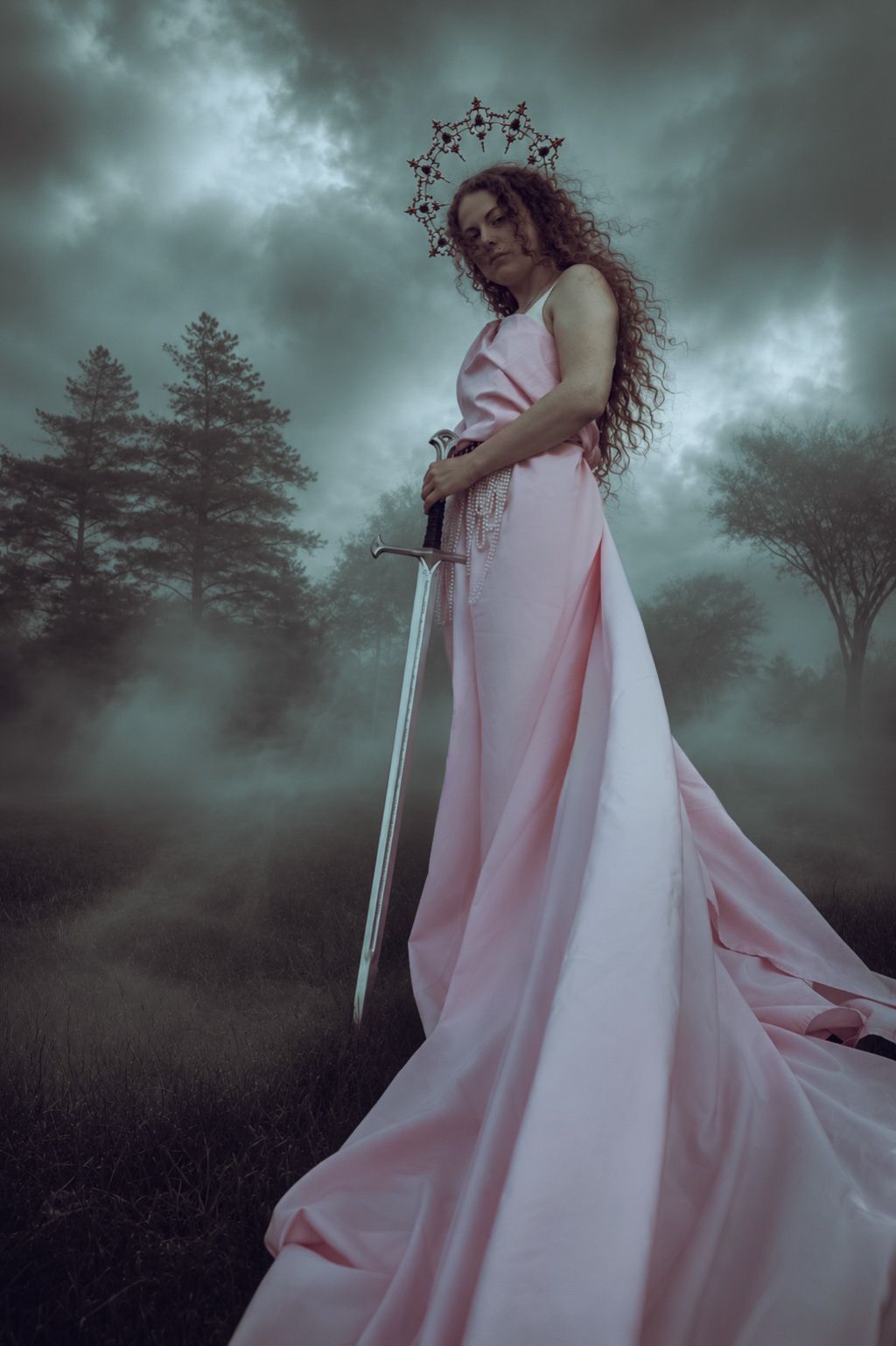 Woman in a flowing pink gown and star crown holds a sword in a misty, moody forest beneath a stormy sky.